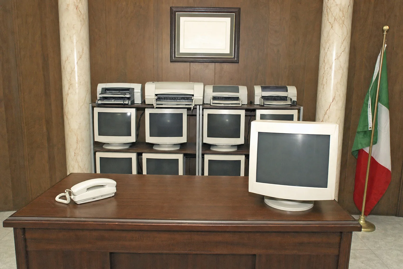 Pekko’s office: a desk, a beige phone, a wall of old monitors and printers, an Italian flag