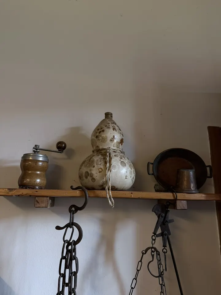 Una mensola con il macinacaffè, una fiasca a forma di zucca e una padella; sotto, le catene del camino