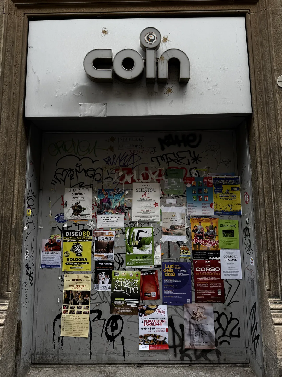 La saracinesca di un ingresso della Coin in via Rizzoli a Bologna: lamiera grigia coperta di graffiti e di volantini, sopra l’insegna «coin» con le lettere a scatola.
